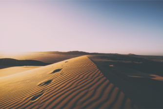 Golden sand dune with visible footprints leading toward the horizon, captured at sunrise or sunset in a wide desert landscape with mountains in the distance and a clear gradient sky transitioning from soft pink to deep blue.